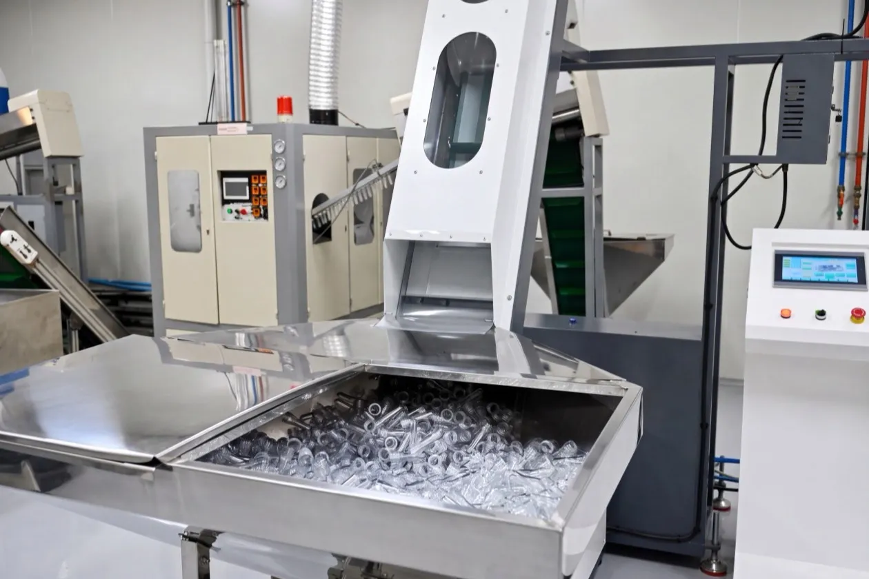 A stainless steel hopper of clear PET preforms feeding an inclined conveyor into a blow-moulding machine.